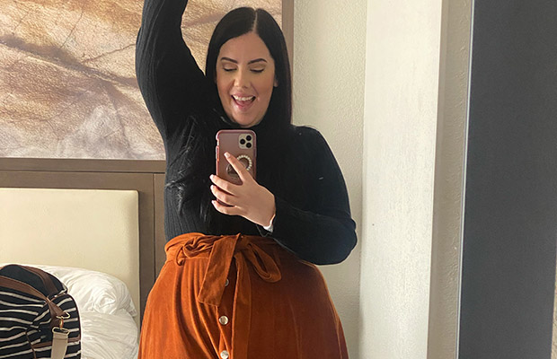 A woman taking a selfie of herself, she has on a long black shirt and a brown skirt