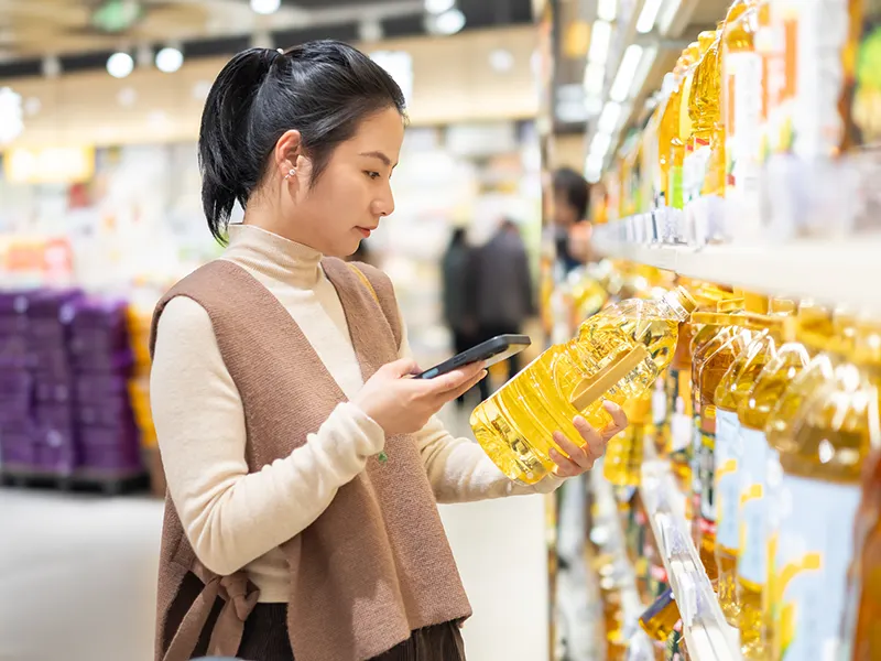 A woman at a grocery store holding an oil bottle with one hand, she holds her phone with the other hand