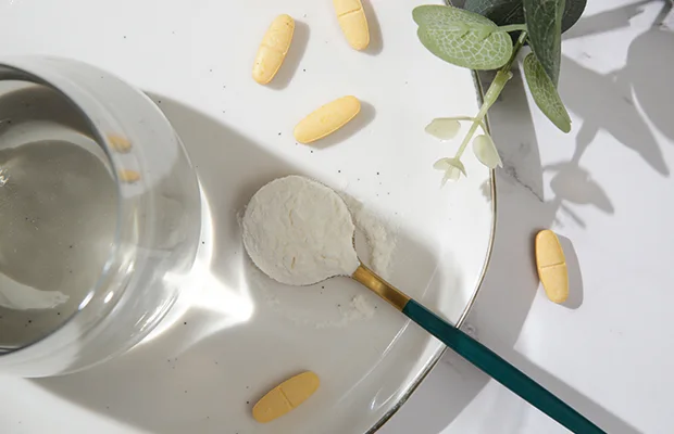 A closeup of a spoon full of collagen powder, there's a glass nearby and some yellow pills