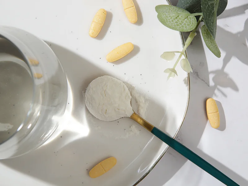 A closeup of a spoon full of collagen powder, there's a glass nearby and some yellow pills