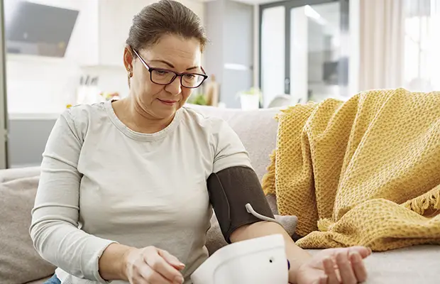 A woman at home sitting on a couch, she is taking her blood pressure with a blood pressure machine