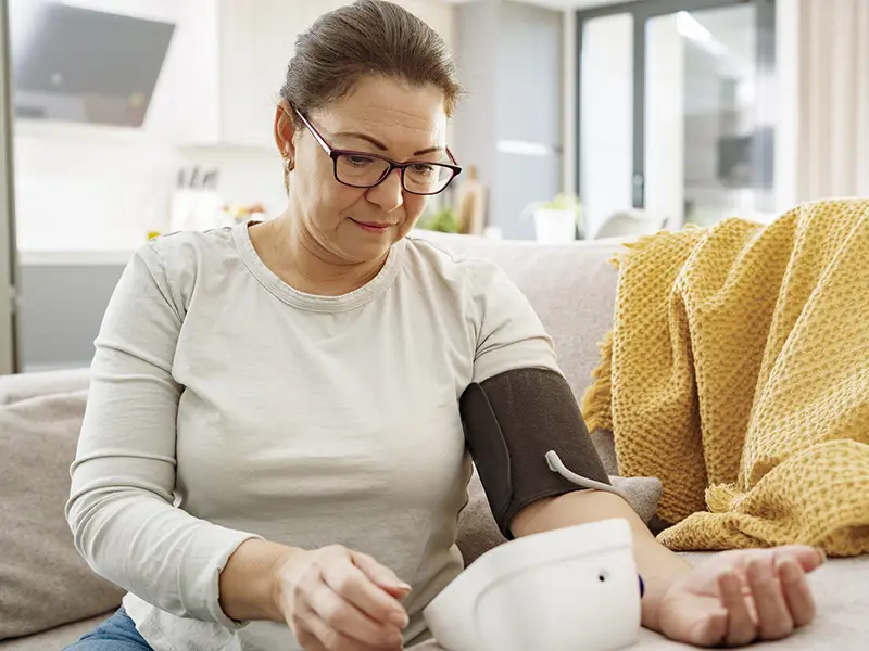 A woman at home sitting on a couch, she is taking her blood pressure with a blood pressure machine
