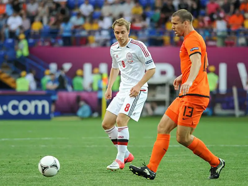Two soccer players competing during a match, one in a white Denmark jersey (number 8) and one in an orange Netherlands jersey (number 13), with a ball on the pitch and stadium crowd in the background.