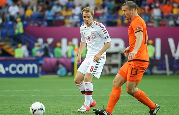 Two soccer players competing during a match, one in a white Denmark jersey (number 8) and one in an orange Netherlands jersey (number 13), with a ball on the pitch and stadium crowd in the background.