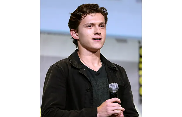 A young man looking forward; he is holding a microphone, is wearing a dark shirt and jacket