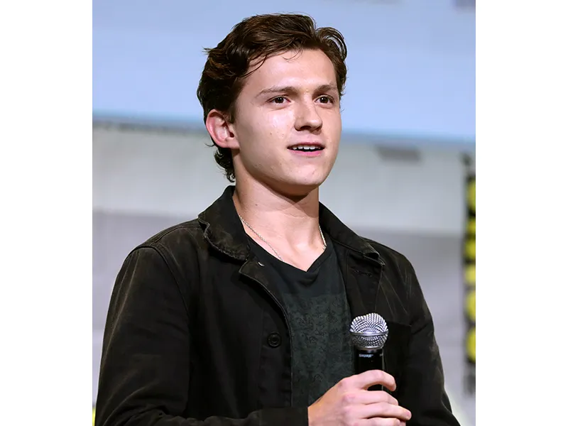 A young man looking forward; he is holding a microphone, is wearing a dark shirt and jacket