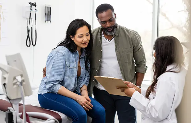 A man and woman sitting together at a doctors office looking over a document a doctor is sharing with them