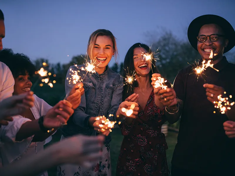 A group of people holding sparklers and smiling, they're outdoors