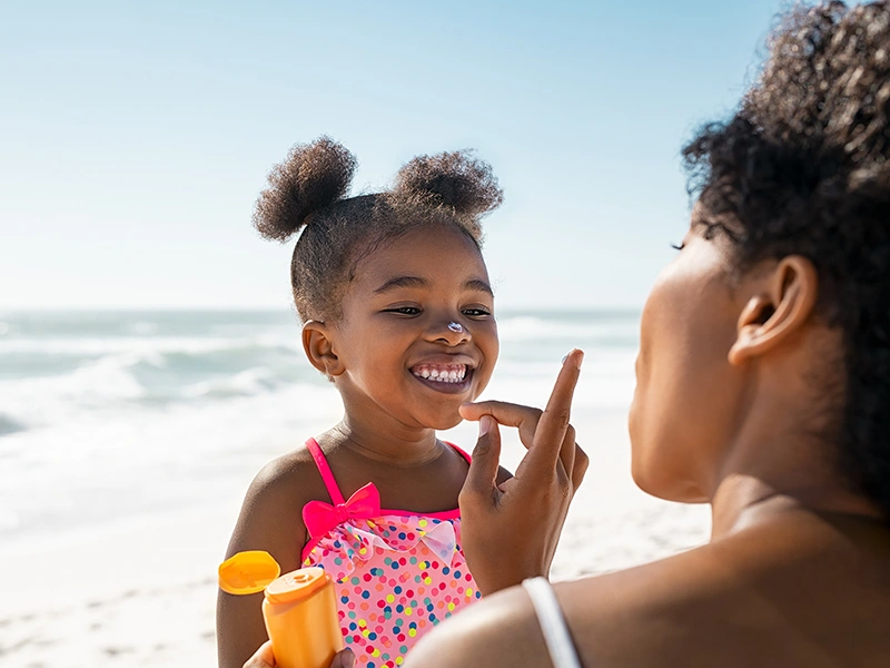 A mom putting on sunscreen on a young girls nose, they are at the beach