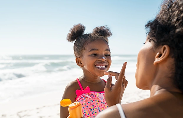 A mom putting on sunscreen on a young girls nose, they are at the beach