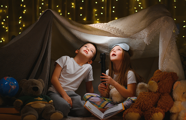 Two children in a fort inside, they have a flashlight and are looking up