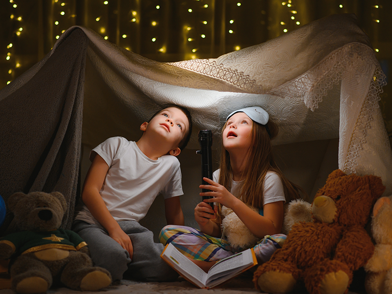 Two children in a fort inside, they have a flashlight and are looking up