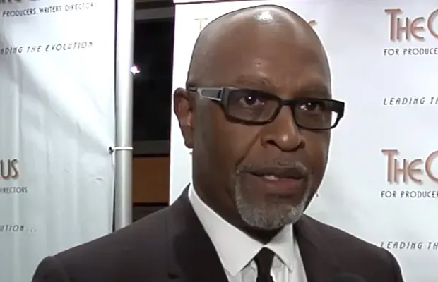 A closeup of a man speaking at a red carpet event, he has on dark glasses and a suit
