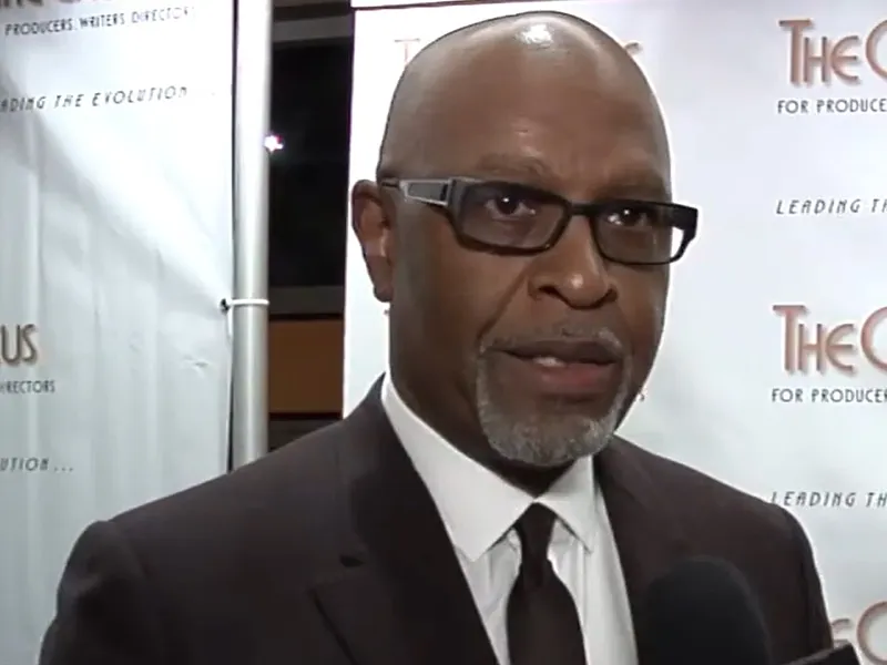A closeup of a man speaking at a red carpet event, he has on dark glasses and a suit