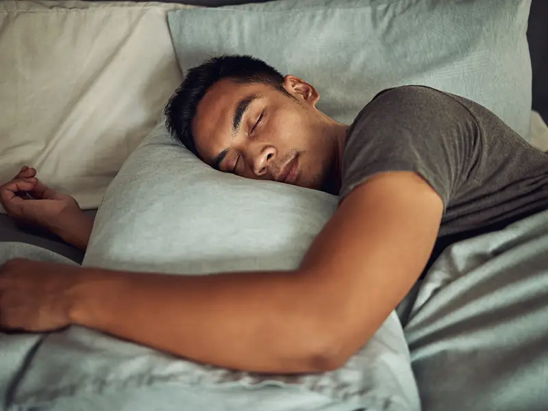 A man sleeping in bed, he has his left hand over his pillow