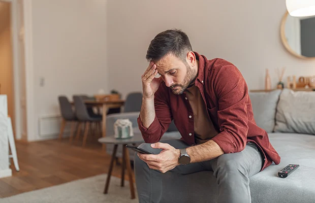 A man sitting on a couch, he is looking down at his phone, his hand is on his forehead