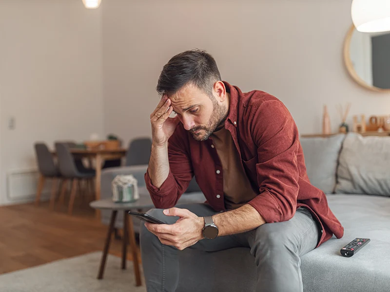A man sitting on a couch, he is looking down at his phone, his hand is on his forehead