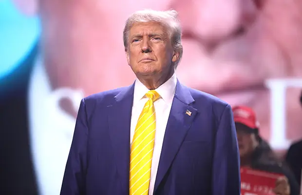 A man standing and looking forward, he has on a dark blue jacket and yellow tie