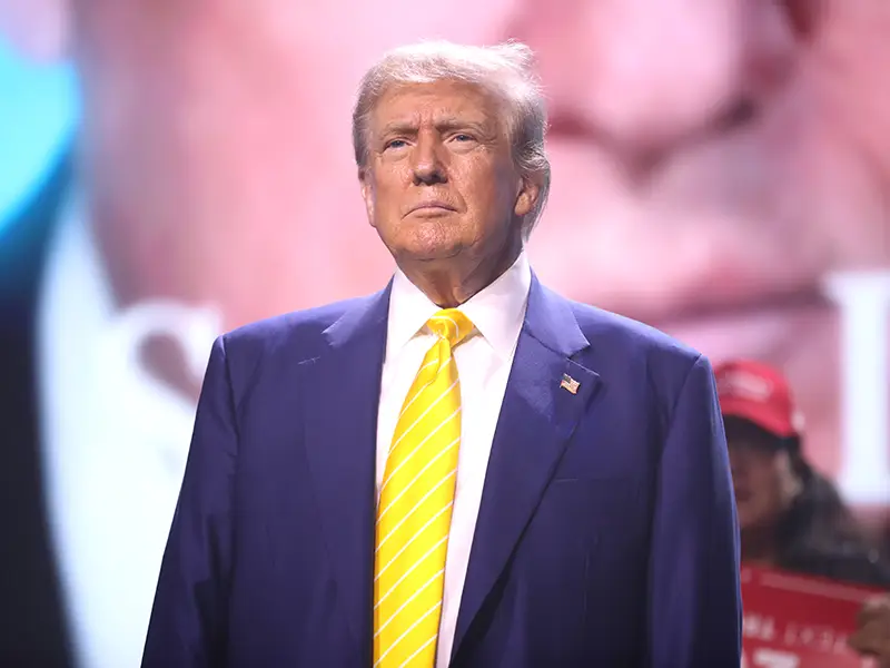 A man standing and looking forward, he has on a dark blue jacket and yellow tie