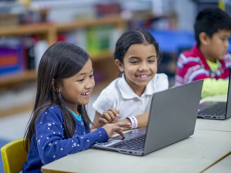 Two young kids smiling, they sit in front of a laptop