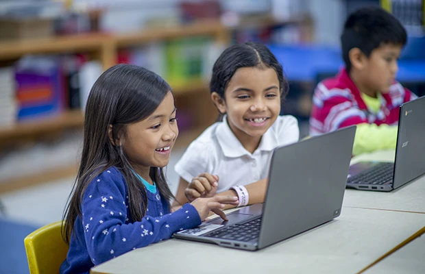 Two young kids smiling, they sit in front of a laptop