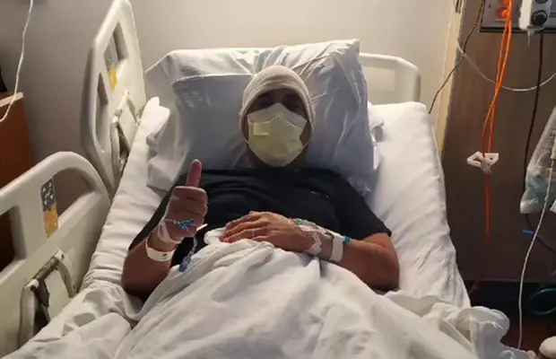 A man in a hospital bed, he has on a mask and head band, he is giving a thumbs up