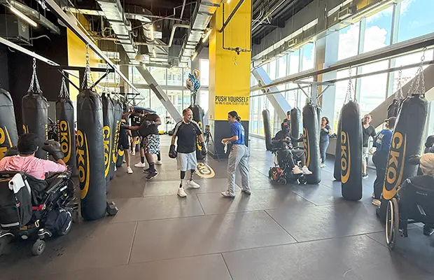Two people practicing kickboxing, there are people around in wheelchairs hitting boxing bags