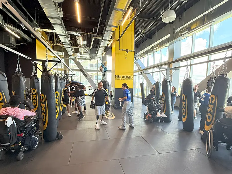 Two people practicing kickboxing, there are people around in wheelchairs hitting boxing bags