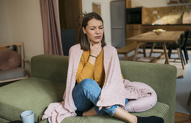A woman sitting on a couch with a blanket wrapped around her shoulders, she is touching her neck as if to indicate that she has throat pain