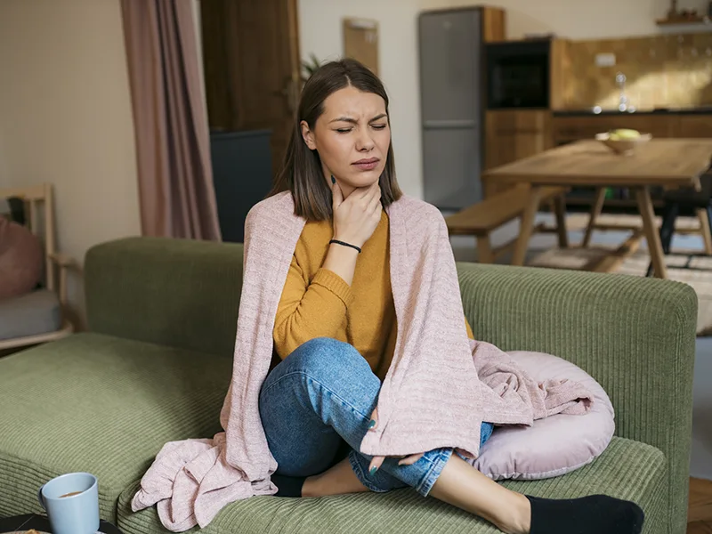 A woman sitting on a couch with a blanket wrapped around her shoulders, she is touching her neck as if to indicate that she has throat pain
