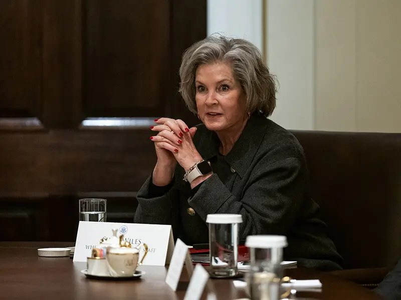 A woman sitting at a table looking out, she has her hands clasped together in front of her, she has on a dark suit jacket