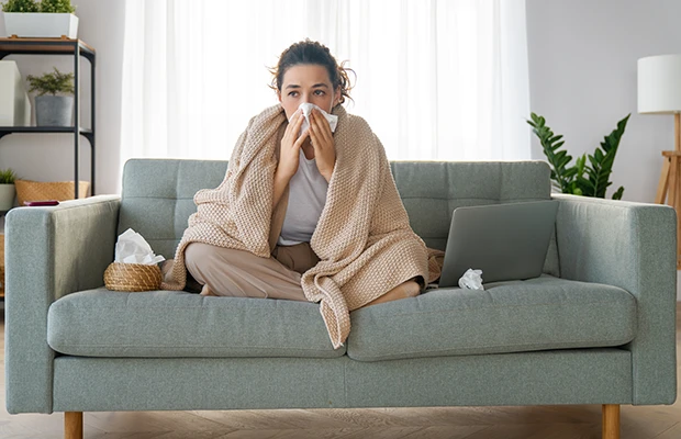 A woman sitting on a couch, she's blowing her nose and has a blanket over her