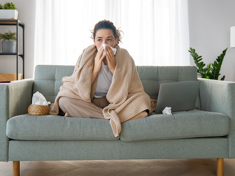 A woman sitting on a couch, she's blowing her nose and has a blanket over her