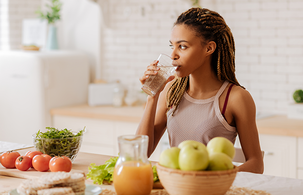 A woman drinking water in her kitchen, there are fruits and vegetables on the counter