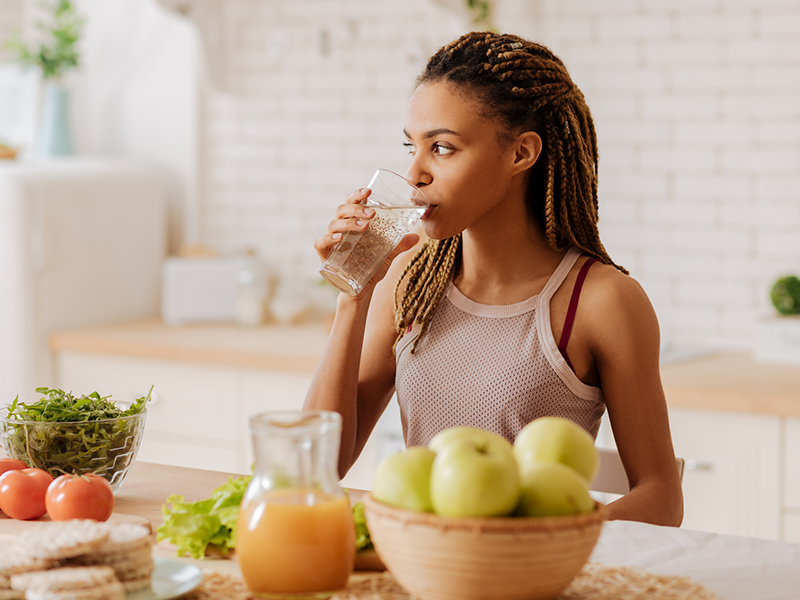 A woman drinking water in her kitchen, there are fruits and vegetables on the counter
