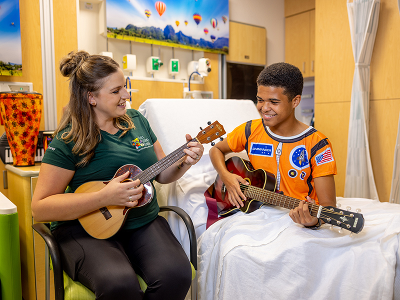 A woman and young boy playing guitars, the young boy is sitting on a medical bed