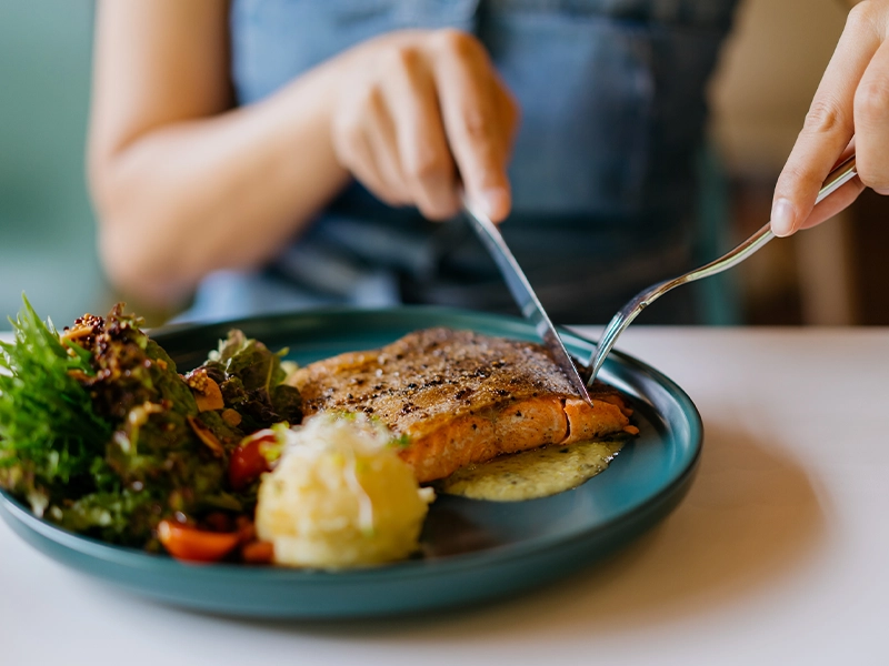 Close-up of a woman's hands cutting a piece of salmon, alongside a salad and mashed potatoes.