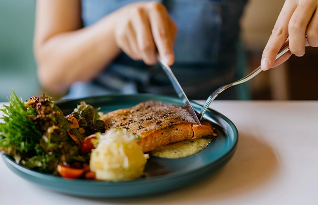 Close-up of a woman's hands cutting a piece of salmon, alongside a salad and mashed potatoes.
