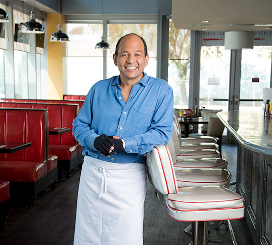 Burn Patient, Ralph Pagano leaning on a chair in a restaurant as he smiles at the camera