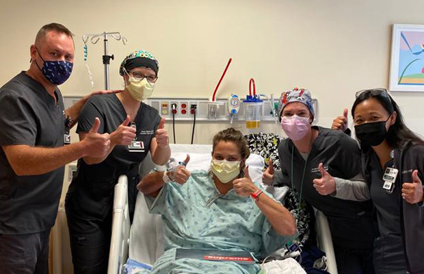 Four medical professionals and a woman who is on a medical bed holding their thumbs up, they all wear face masks