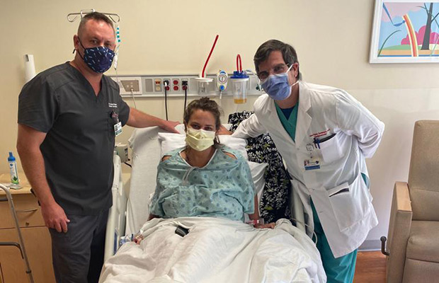 Two medical professionals taking a picture with a patient who is laying on a hospital bed, they all wear face masks
