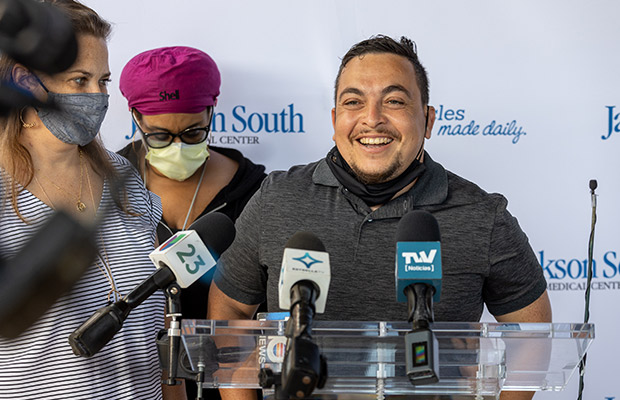 A middle aged man speaking to the media, he is standing next to two people who are wearing face masks