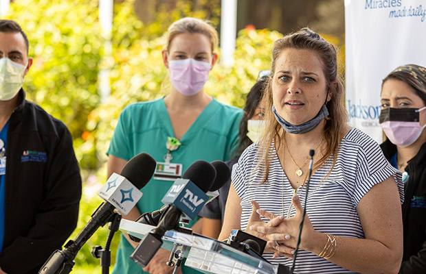 A woman at a press event speaking to the media, there are medical professionals standing behind her