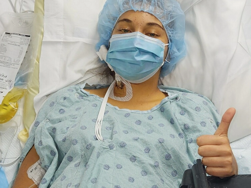 A woman giving a thumbs-up while lying on a hospital gurney, wearing a cap and a surgical mask.