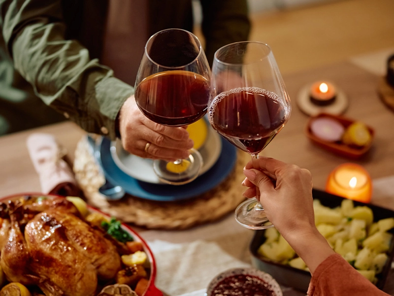 Close-up of two people toasting with glasses of red wine next to a table full of Christmas dishes
