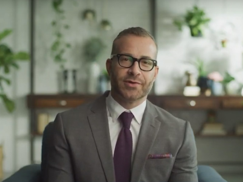 A man smiling at the camera, he is wearing a gray suit, purple tie, and white collar shirt