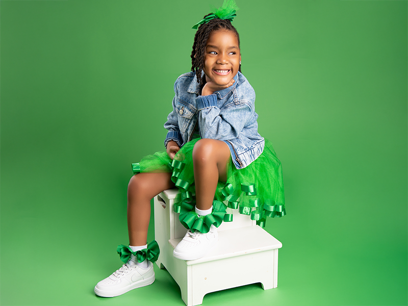 Little girl dressed in green sitting on a small white ladder, smiling and looking to the side.