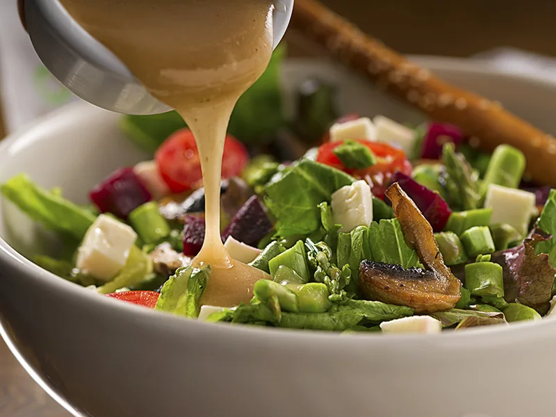 A closeup of salad dressing being pored over a bed of lettuce and vegetables