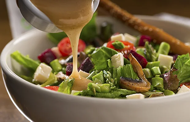 A closeup of salad dressing being pored over a bed of lettuce and vegetables