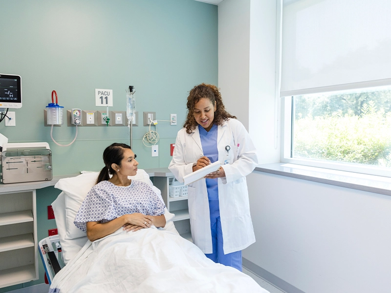 A patient laying on a hospital bed, there's a medical professional speaking with her and showing her something on a clipboard